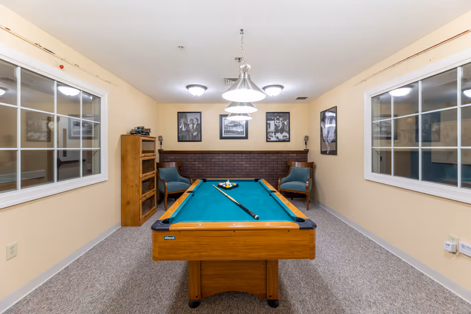 A well-lit game room with a pool table in the center, featuring a green felt surface and a cue stick resting on it. The room has beige walls, two large windows on either side, a wooden cabinet, and two blue upholstered chairs against a brick accent wall. Three pendant lights hang above the pool table, and framed black-and-white photographs are displayed on the walls.
