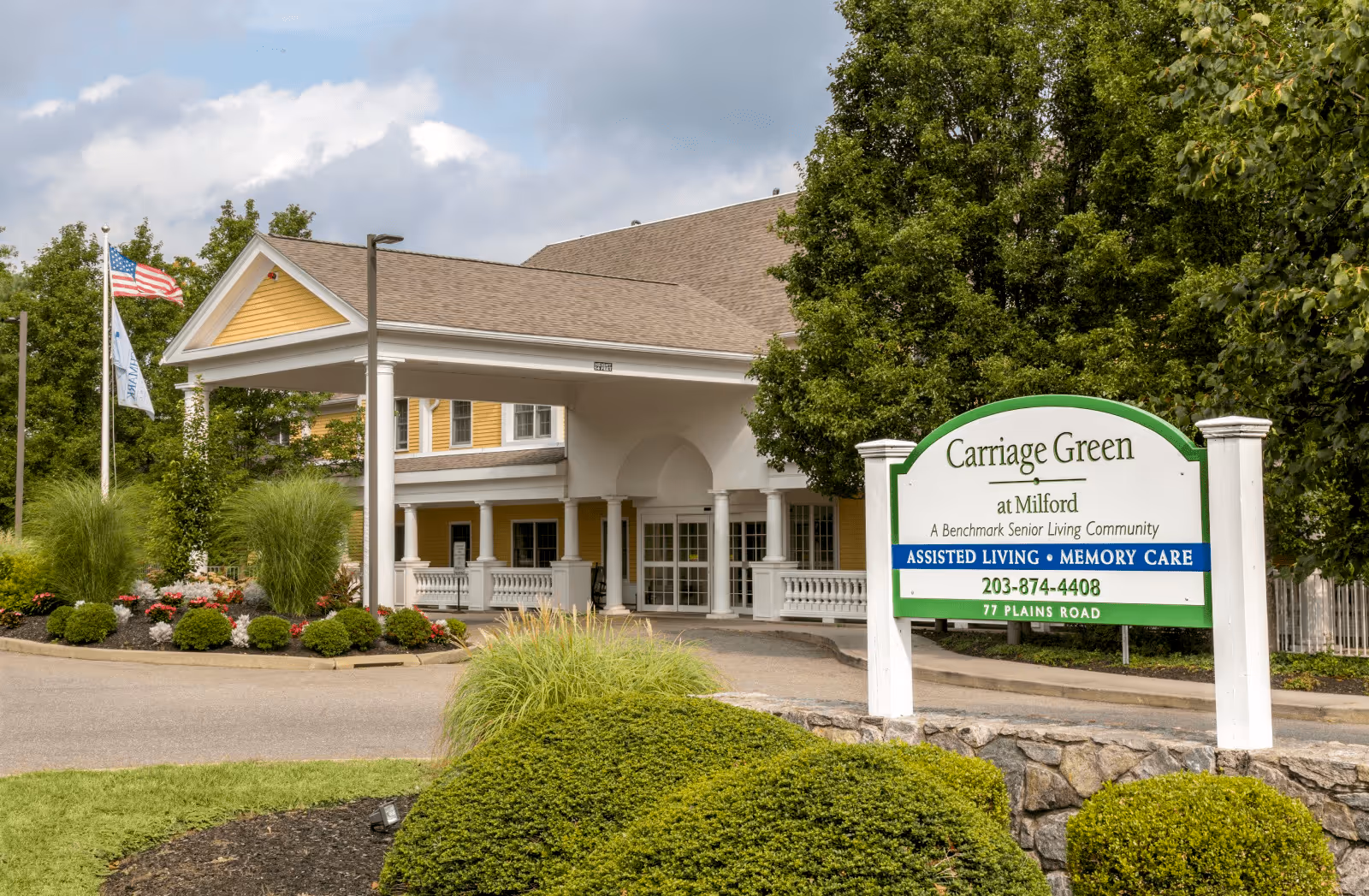 Exterior view of Carriage Green at Milford senior living community entrance with a covered driveway, landscaped bushes, flowers, and two flagpoles with flags.