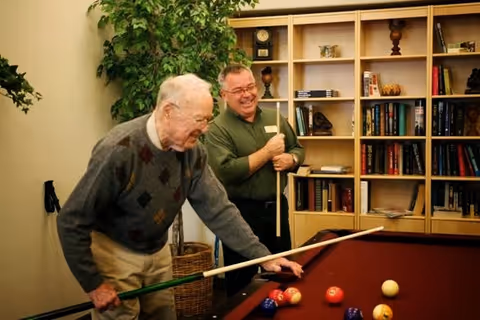 An elderly man and a middle-aged man playing pool together in a room with a bookshelf filled with books and decorative items in the background. The elderly man is leaning over the pool table preparing to take a shot, while the middle-aged man stands behind him smiling and holding a pool cue.