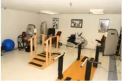 A therapy room in Haida Manor-Therapy Department featuring exercise equipment including parallel bars for walking practice, stationary bikes, and other rehabilitation machines. The room has white walls with framed pictures and bright overhead lighting.