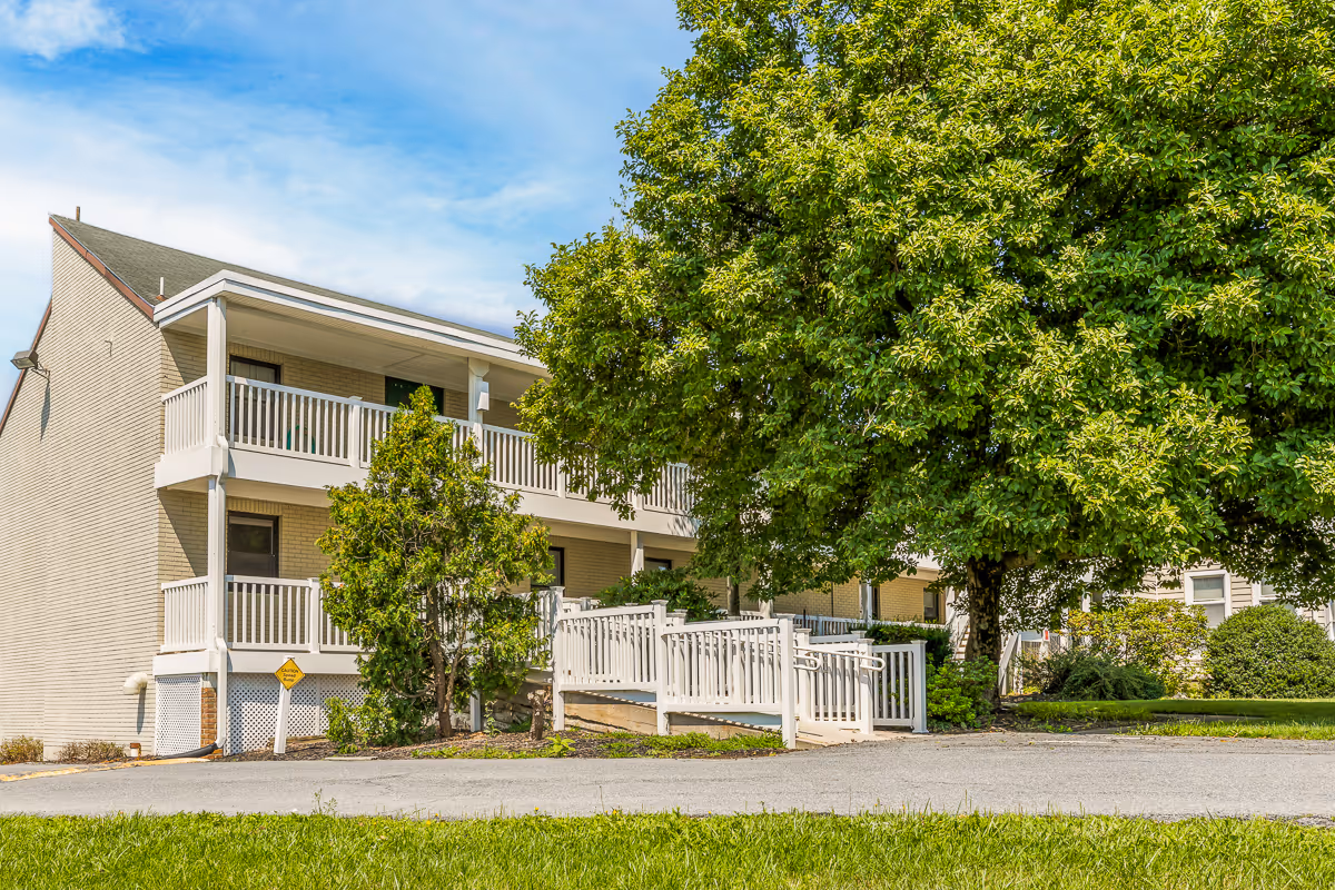 Two-story light-brick senior living building with white balconies and an entry ramp next to a large leafy tree.