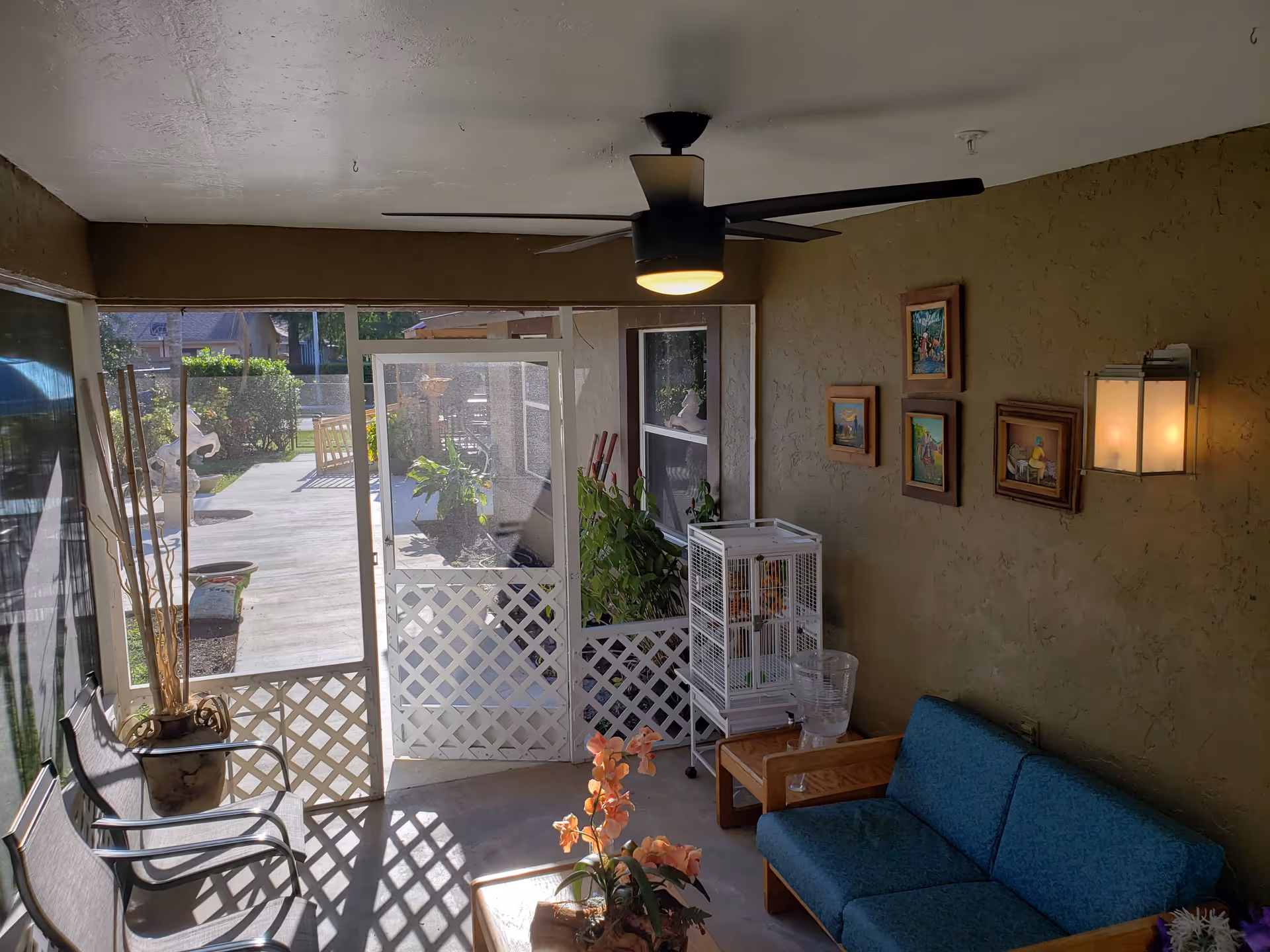 A cozy enclosed patio area with a blue cushioned wooden sofa, a small wooden table with a flower arrangement, a white birdcage on a stand, and two metal chairs. The walls are textured and painted brown, decorated with four framed paintings and a wall-mounted light fixture. A ceiling fan with a light is mounted on the white ceiling. The patio opens to an outdoor walkway with greenery and garden decorations visible outside.