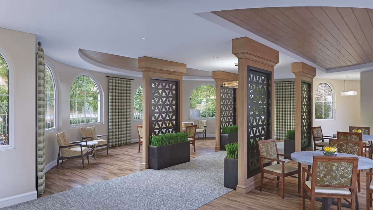 A bright and spacious common area with large arched windows, patterned curtains, wooden flooring, and a carpeted walkway. The room features wooden columns with decorative panels and planters with green plants. There are several round tables with floral upholstered chairs and small seating areas with armchairs and side tables near the windows.