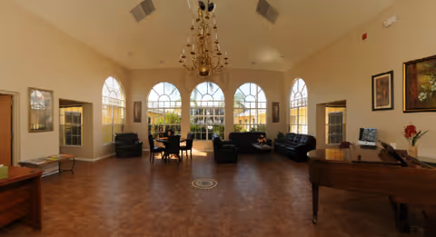 Spacious assisted-living common room with large arched windows, a chandelier, sofas, a piano, and a central seating area.
