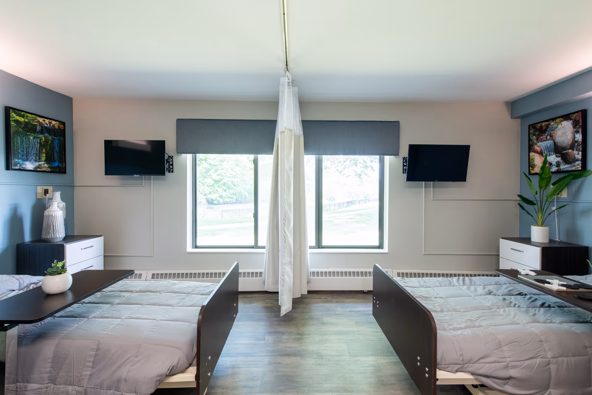 Shared resident bedroom with two hospital-style beds separated by a central privacy curtain, TVs on the walls, windows, and bedside dressers.