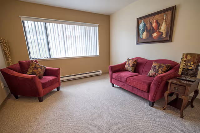 A small living room with a maroon loveseat and matching armchair, both with decorative pillows. There is a wooden side table with a decorative lamp next to the loveseat. A large window with vertical blinds lets in natural light, and a framed painting of vases hangs on the beige wall above the loveseat.