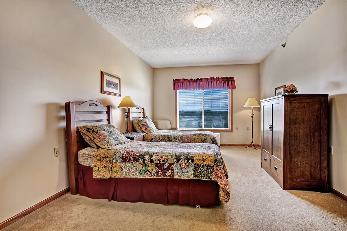 A bedroom with two twin beds featuring floral patchwork quilts, a wooden nightstand with a lamp between the beds, a wooden armoire, a floor lamp, and a window with a red valance letting in natural light.