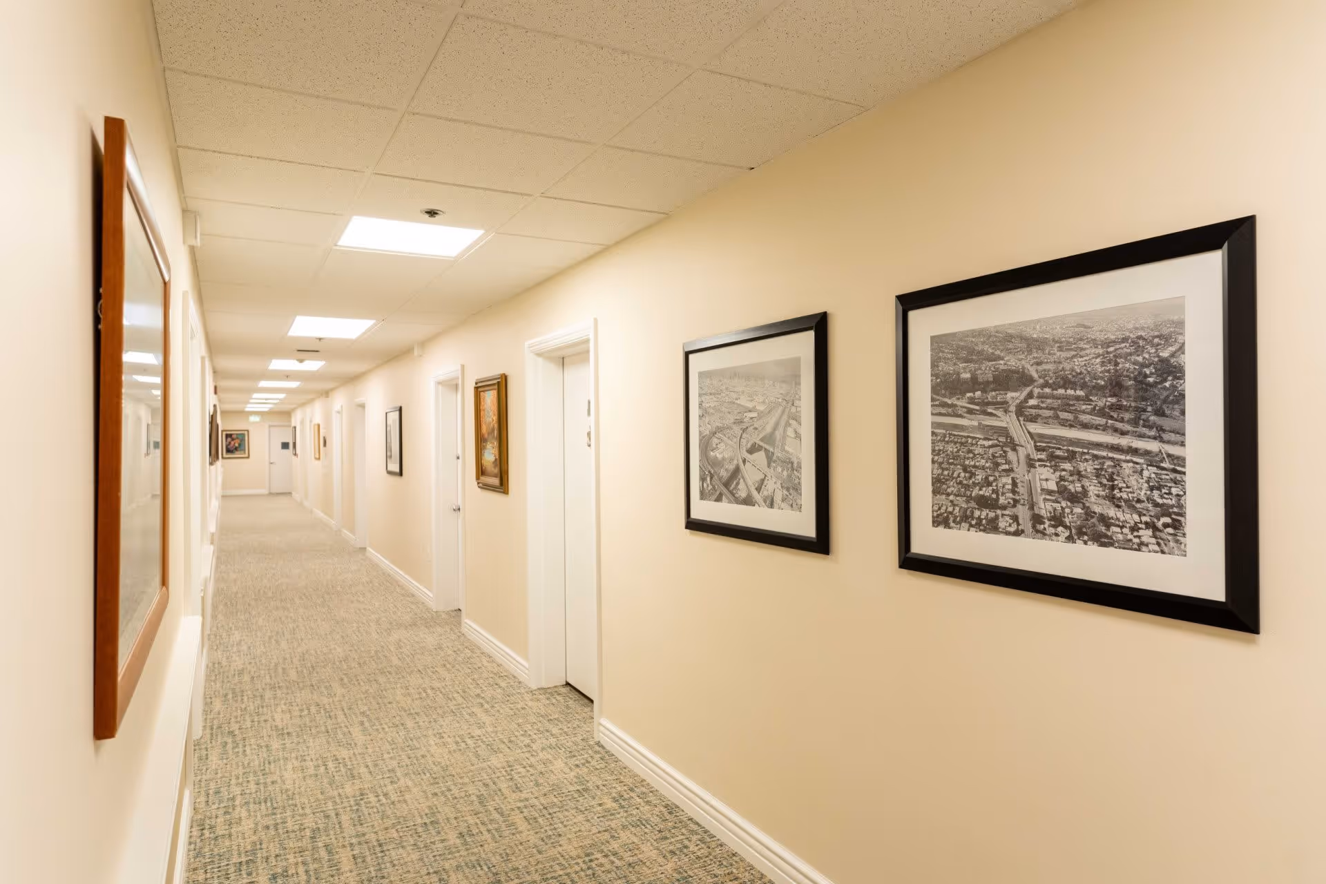 A long, well-lit hallway in a senior living facility with beige walls and carpeted floor. Several framed pictures and artworks hang on the walls. Multiple white doors line the hallway, and the ceiling has recessed lighting panels.