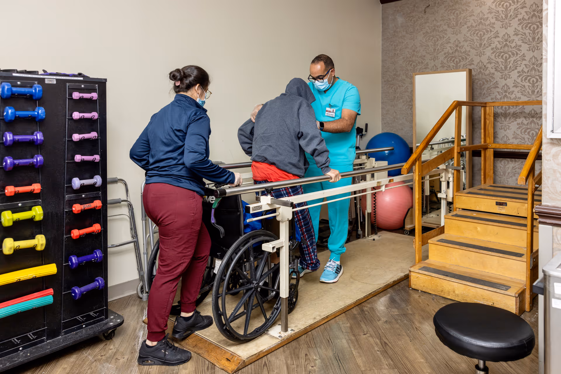 A physical therapy session in a rehabilitation center where a patient wearing a hoodie and plaid pants is assisted by two healthcare workers, one in teal scrubs and the other in casual attire, as the patient practices walking with parallel bars. The room contains exercise equipment including colorful dumbbells, exercise balls, and a wooden staircase for mobility training.