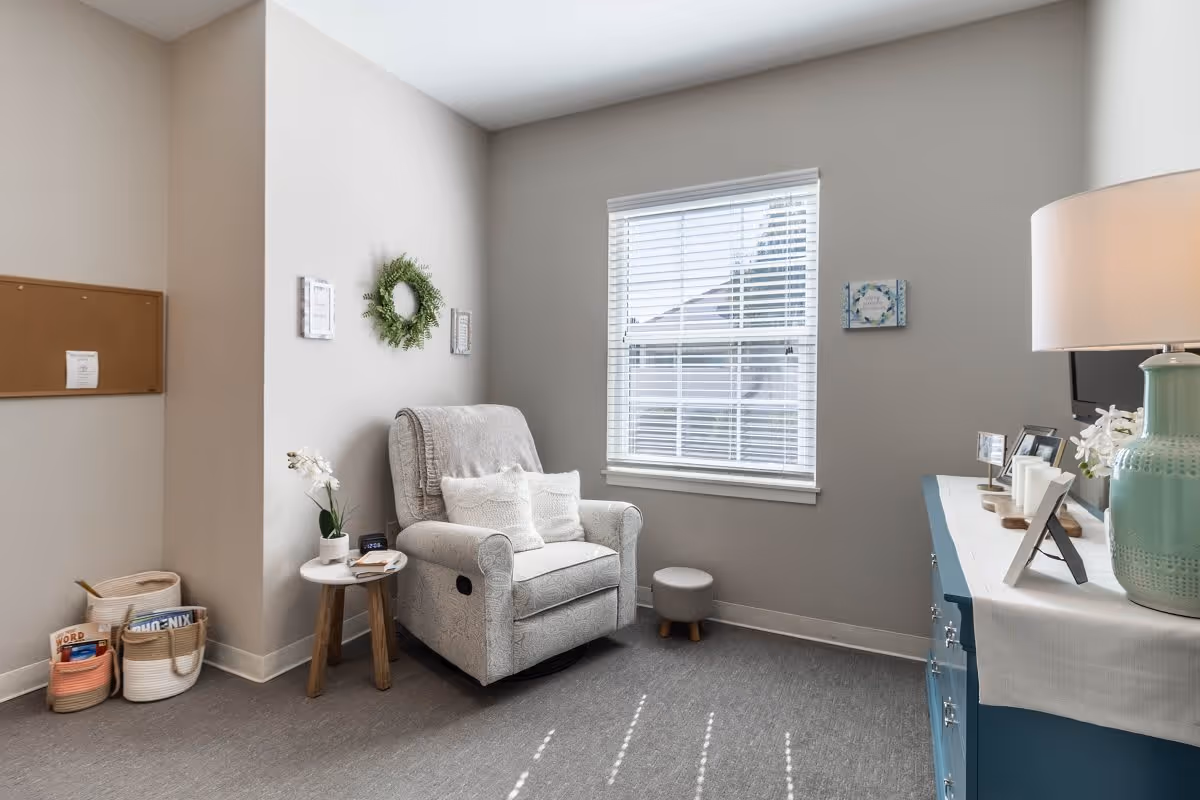 A cozy interior room with a comfortable gray armchair adorned with two white pillows, positioned next to a small round wooden side table holding a white orchid plant and a clock. The room has light gray walls with a window featuring white blinds letting in natural light. A blue dresser with a white runner, a green lamp, and framed photos is on the right side. On the left, there is a corkboard on the wall and baskets on the floor containing magazines and other items. The floor is covered with gray carpet.