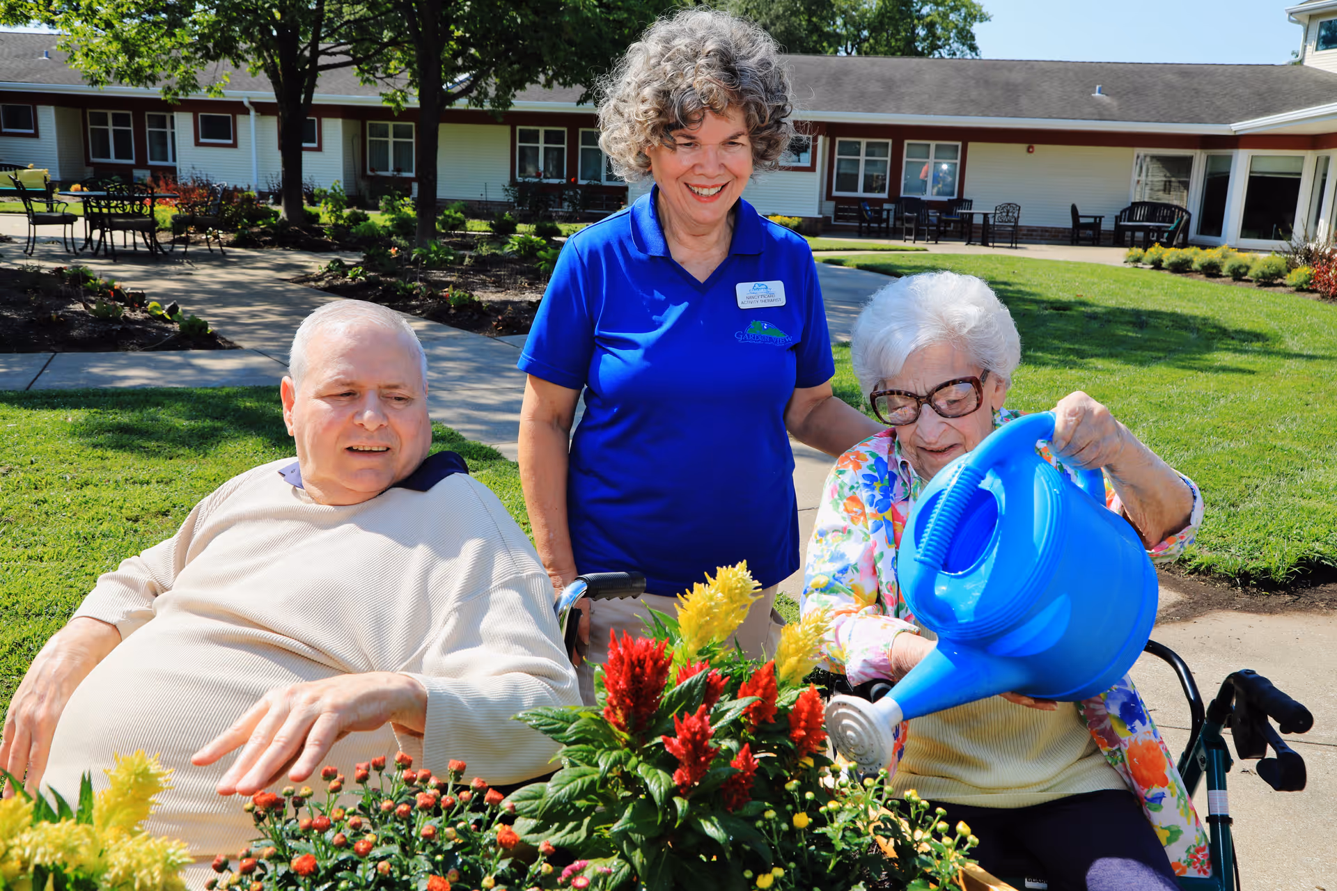 An elderly woman in a wheelchair waters colorful flowers with a blue watering can while a man sits nearby and a smiling caregiver in a blue shirt stands behind them in a sunny garden area outside a senior living facility.