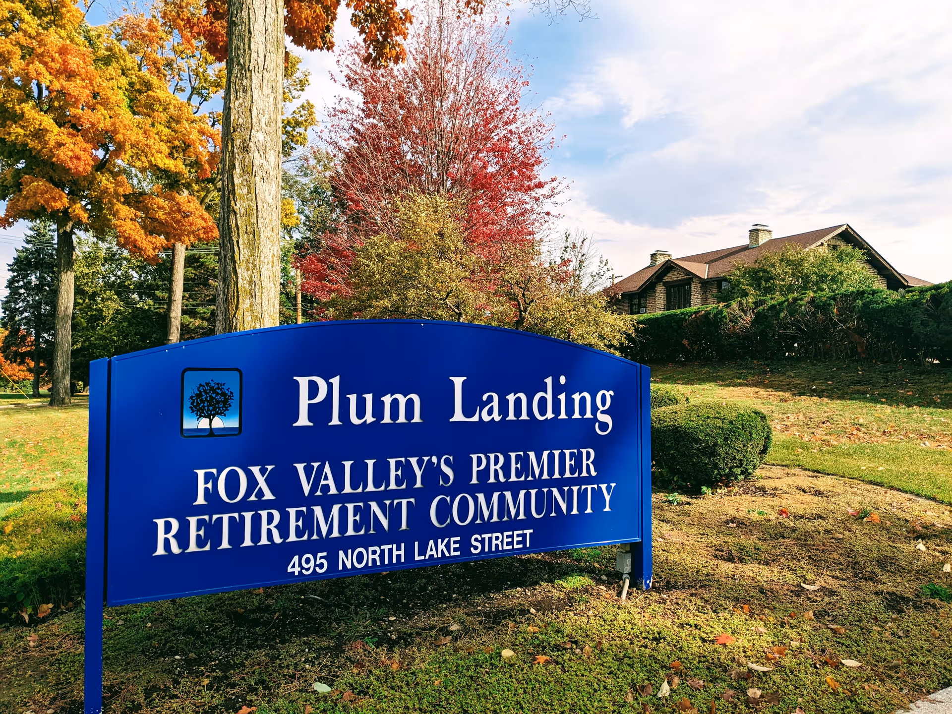 A blue sign for Plum Landing, Fox Valley's premier retirement community, located at 495 North Lake Street. The sign is set outdoors with autumn trees displaying vibrant orange, red, and yellow leaves, and a building partially visible in the background.