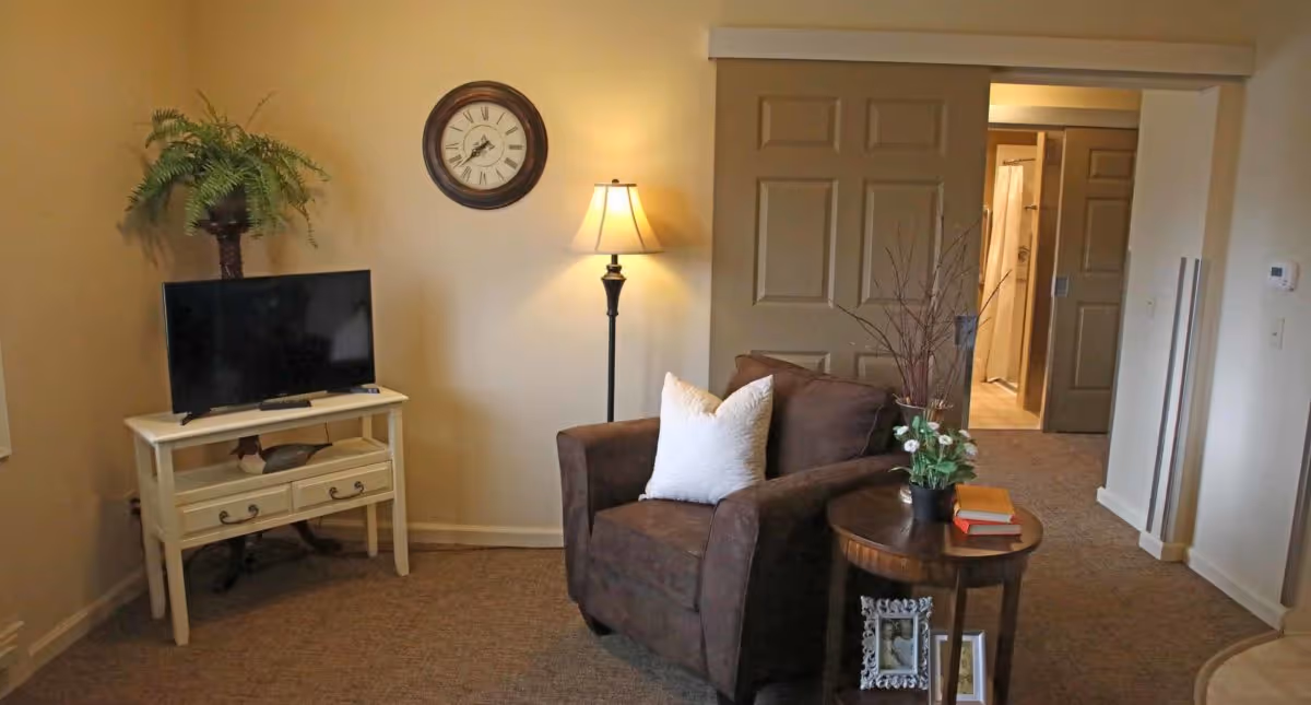 A cozy living room area featuring a brown armchair with a white pillow, a round wooden side table with a small plant and books, a floor lamp, a wall clock, and a TV on a white stand with drawers. The room has beige walls and carpeted flooring, with a hallway leading to another room visible in the background.