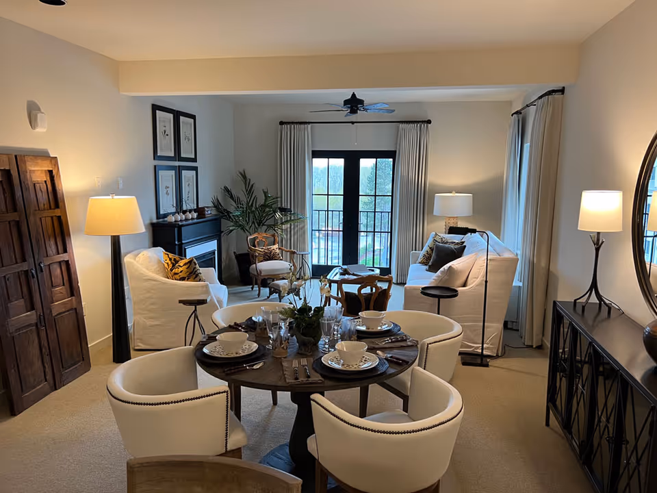 A cozy and well-lit living and dining area featuring a round dining table set with dishes and glassware surrounded by four white upholstered chairs. The living area includes a white sofa, an armchair, a small wooden chair, a fireplace, and several lamps providing warm lighting. Large windows with curtains and a glass door offer a view of the outdoor area.