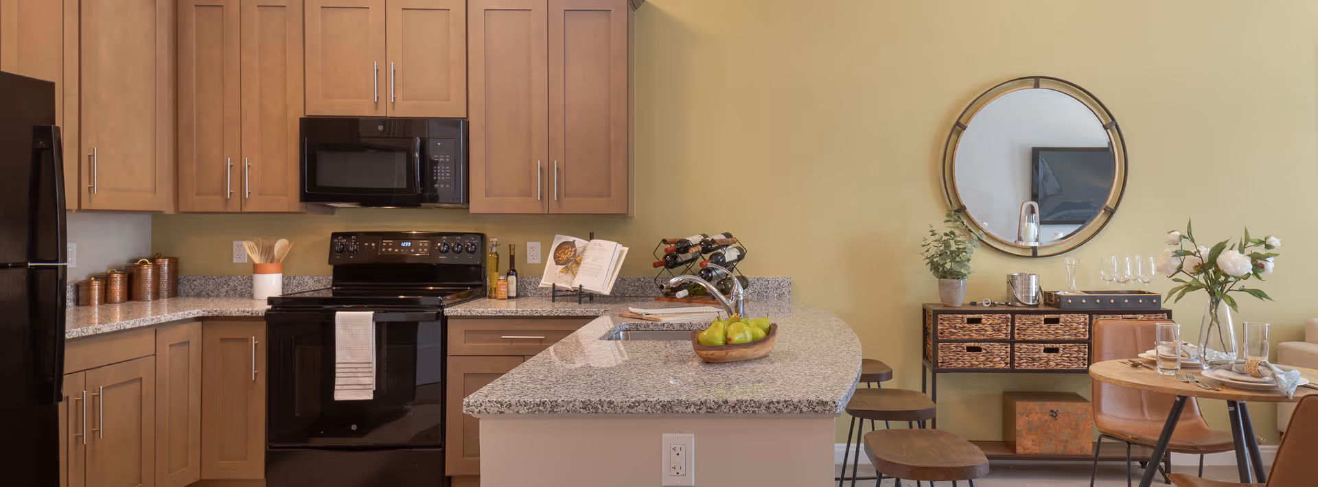 Modern kitchen with light wood cabinets, black appliances including a refrigerator, stove, and microwave. Granite countertops with a kitchen island featuring a sink, a bowl of green apples, and a wine rack. Adjacent dining area with a round wooden table, brown leather chairs, a sideboard with woven drawers, a round mirror on the wall, and decorative plants and glassware.
