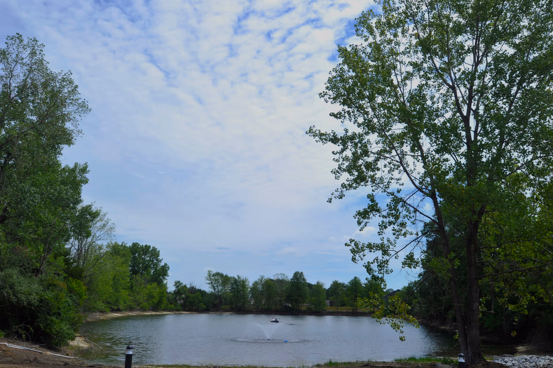 A peaceful outdoor scene featuring a small lake surrounded by green trees under a partly cloudy sky. There is a fountain in the middle of the lake and a person in a small boat. The area appears calm and natural.