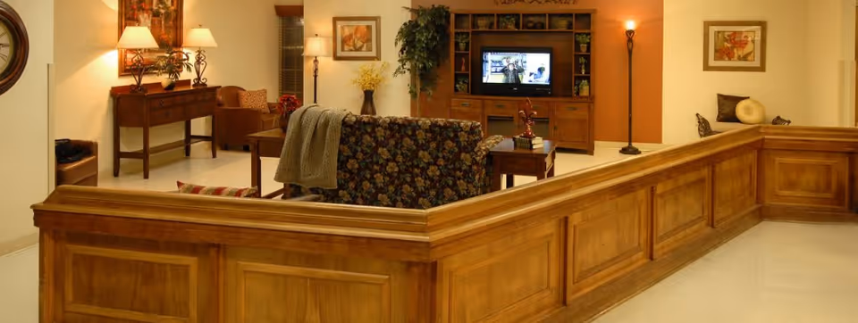 A cozy living room area with wooden paneling and furniture, including a floral-patterned sofa, a wooden coffee table, a TV on a wooden entertainment center, lamps, framed artwork on the walls, and decorative plants.