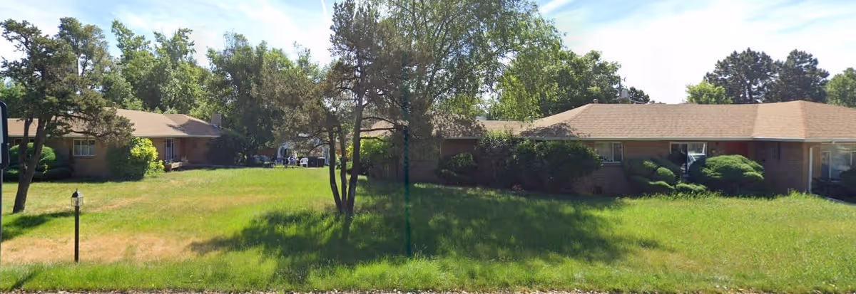 A single-story brick building surrounded by green grass and trees under a blue sky with some clouds. The building appears to be part of a residential or assisted living facility with windows and bushes along the front.