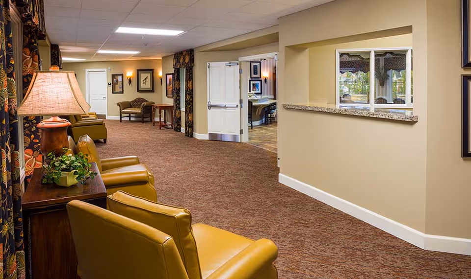 Carpeted interior lounge/hallway with yellow chairs, side tables, a lamp and a reception window in a memory care community.