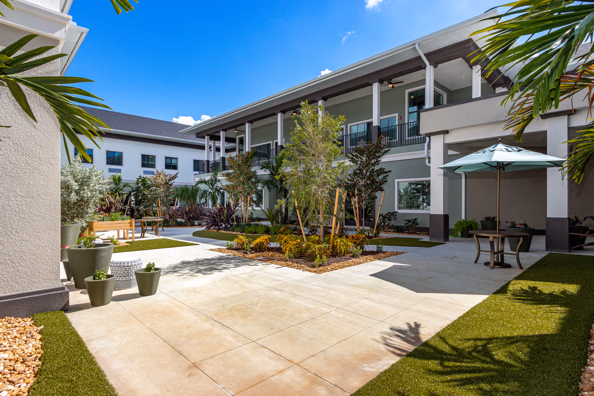 Outdoor courtyard area at Atrium at Liberty Park with paved walkways, green artificial turf, potted plants, small trees, and seating areas including a table with an umbrella. The building surrounding the courtyard has two stories with balconies and windows under a clear blue sky.