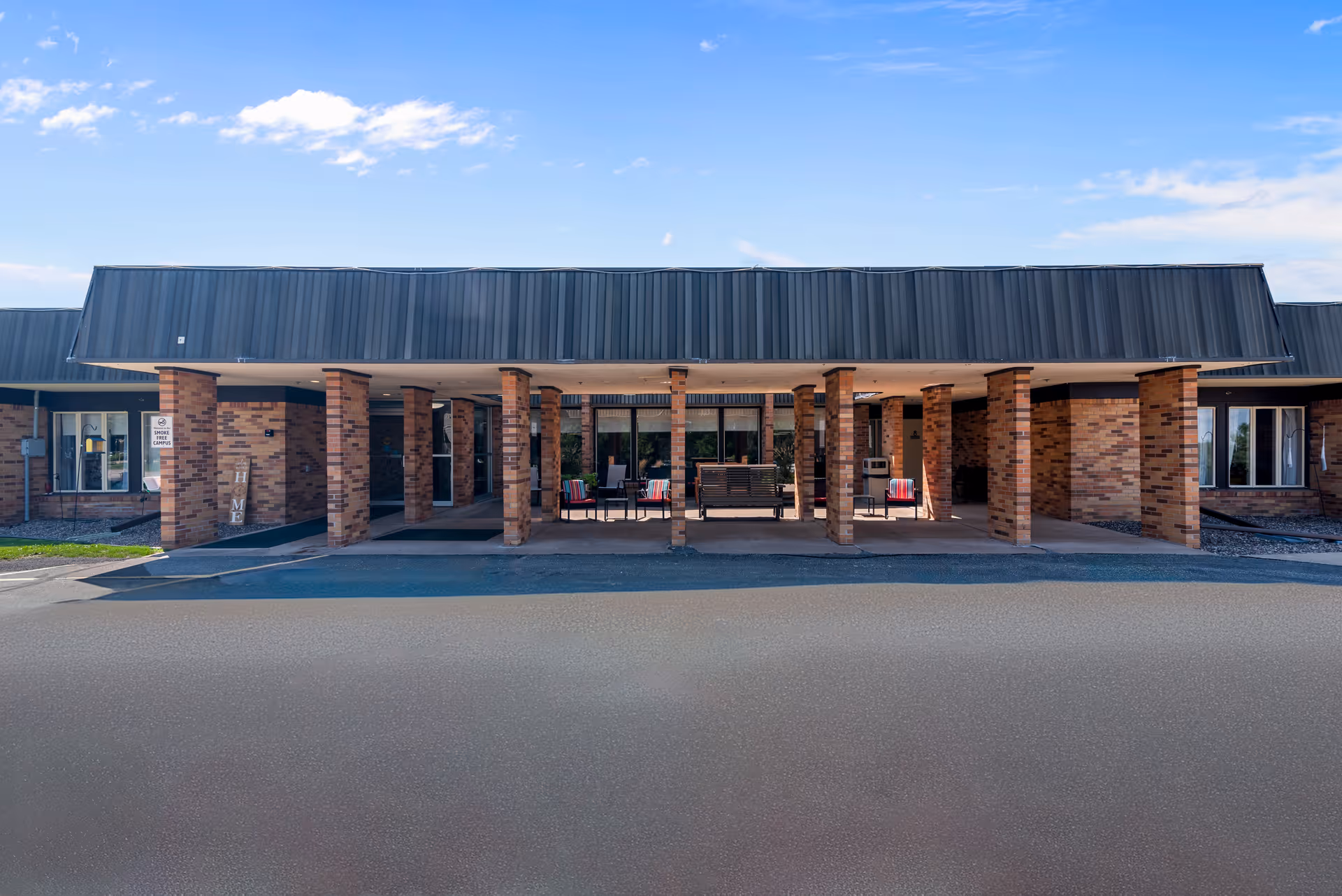 Front exterior view of a single-story brick building with a covered entrance supported by multiple brick columns. There are chairs and a bench under the covered area, and the sky is clear with a few clouds.