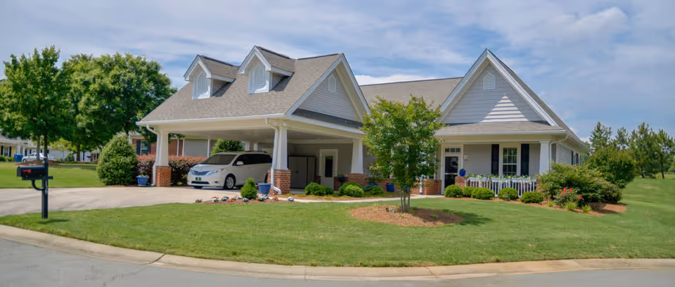 Single-story light-gray building with a covered carport, a white car parked underneath, and a manicured lawn and landscaping in front.