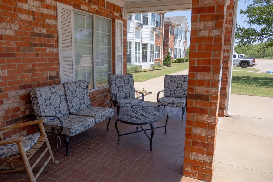 Covered outdoor patio area with brick walls and tiled floor, featuring cushioned metal chairs and a matching round metal table. A wooden rocking chair is also visible. In the background, there is a view of a parking lot, grassy area, and a multi-story building with white siding and brick accents.