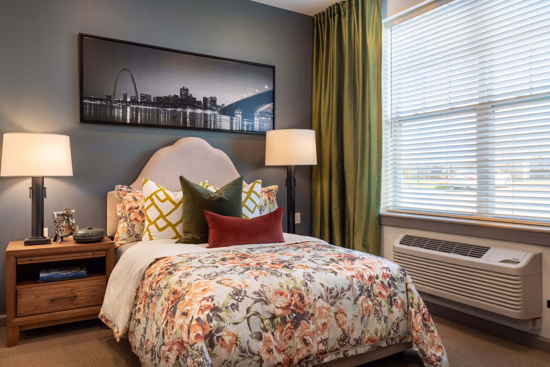A cozy bedroom with a floral patterned bedspread on a bed with multiple decorative pillows. There are two bedside tables each with a lamp, and a black and white cityscape photo hanging above the bed. A window with white blinds and green curtains is on the right side, with an air conditioning unit below it.