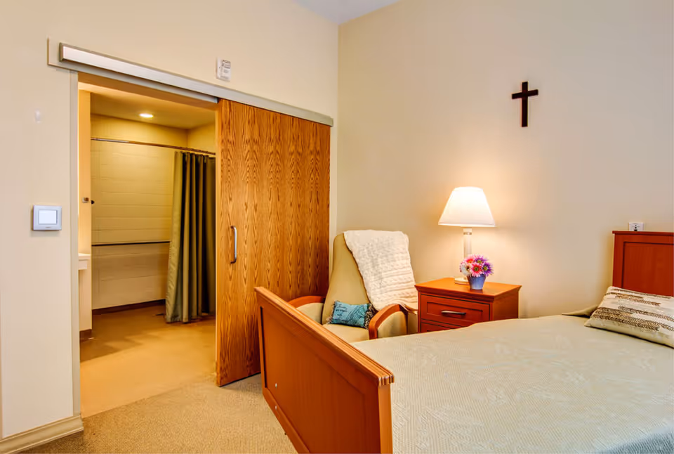 A cozy bedroom in a senior living facility featuring a wooden bed with a beige bedspread, a wooden nightstand with a lamp and a small vase of flowers, a comfortable armchair with a white throw blanket and a blue pillow, and a wooden sliding door leading to a bathroom with a shower curtain. A cross is mounted on the wall above the nightstand.