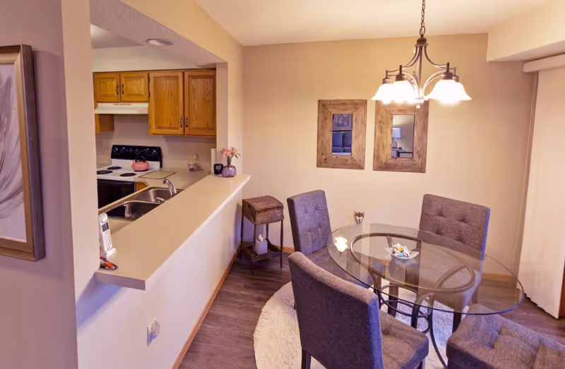 Interior view of a senior living facility dining area with a round glass table surrounded by four gray upholstered chairs on a circular rug. A small side table with a basket and decorative items is against the wall, which has two wooden framed mirrors. To the left, a kitchen area with wooden cabinets, a stove, and a sink is visible. A hanging light fixture with multiple bulbs illuminates the dining area.