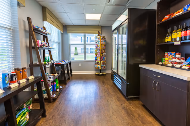 A small convenience store area inside a senior living facility with wooden shelves stocked with snacks, beverages, and personal care items. There is a large window with blinds and a display rack with greeting cards near the back wall. The floor is wood, and the walls are painted light gray.