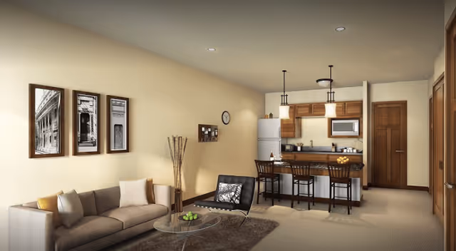 A cozy senior living interior featuring a living room with a beige sofa, a black chair, a glass coffee table with green apples, and three framed black-and-white photos on the wall. Adjacent to the living room is a kitchen area with wooden cabinets, a refrigerator, microwave, and a breakfast bar with three chairs. The walls are painted light beige, and the floor is carpeted.