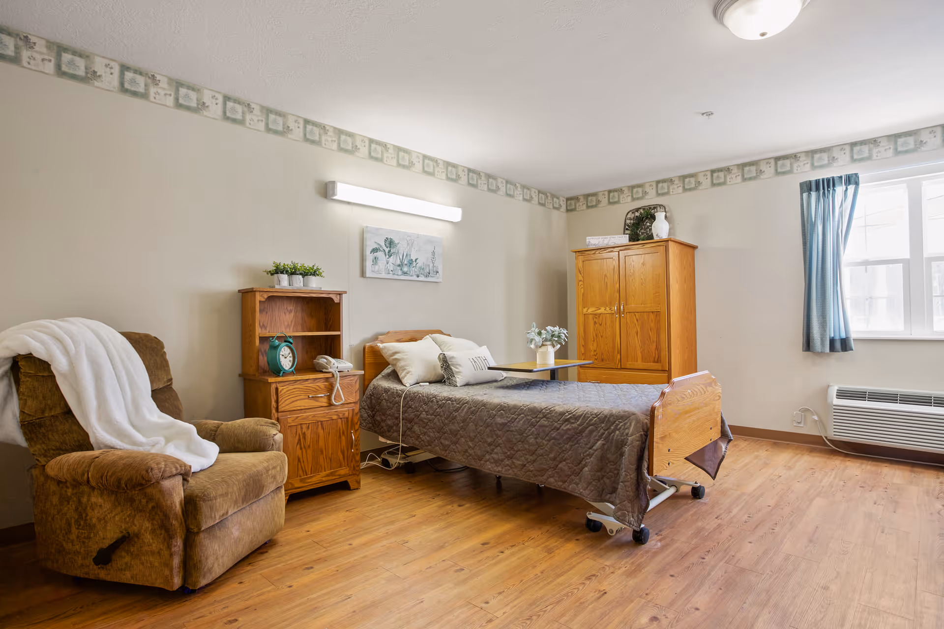 A senior living bedroom with a single bed covered in a gray quilt, a wooden nightstand with a green clock and a phone, a brown recliner chair with a white blanket draped over it, a wooden wardrobe, a small table with a plant, and a window with blue curtains letting in natural light.