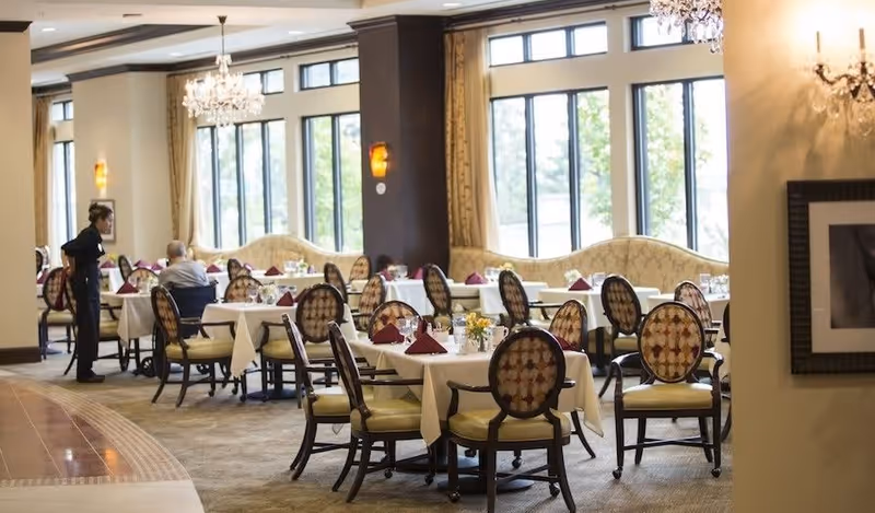 A spacious dining room with multiple tables covered in white tablecloths, each set with folded burgundy napkins and small flower arrangements. The room features large windows letting in natural light, elegant chandeliers, and upholstered chairs with patterned backs. A staff member is attending to seated guests.