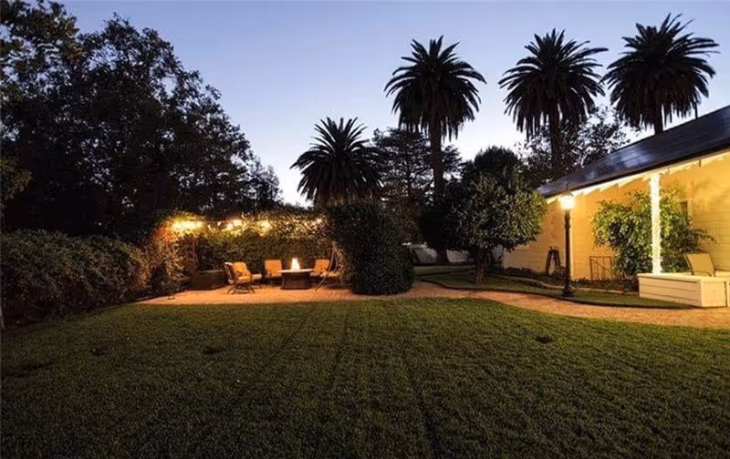 Outdoor garden area at dusk with a well-maintained lawn, palm trees, and a seating area with chairs around a fire pit. The area is softly lit with string lights and a lamp post near a building with yellow walls and white trim.