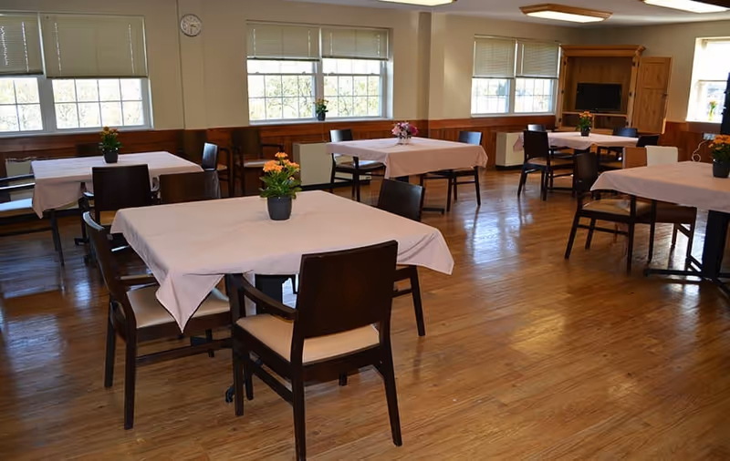 A bright dining room with several square tables covered in light tablecloths, wooden chairs, small potted plants on each table, and large windows letting in daylight.