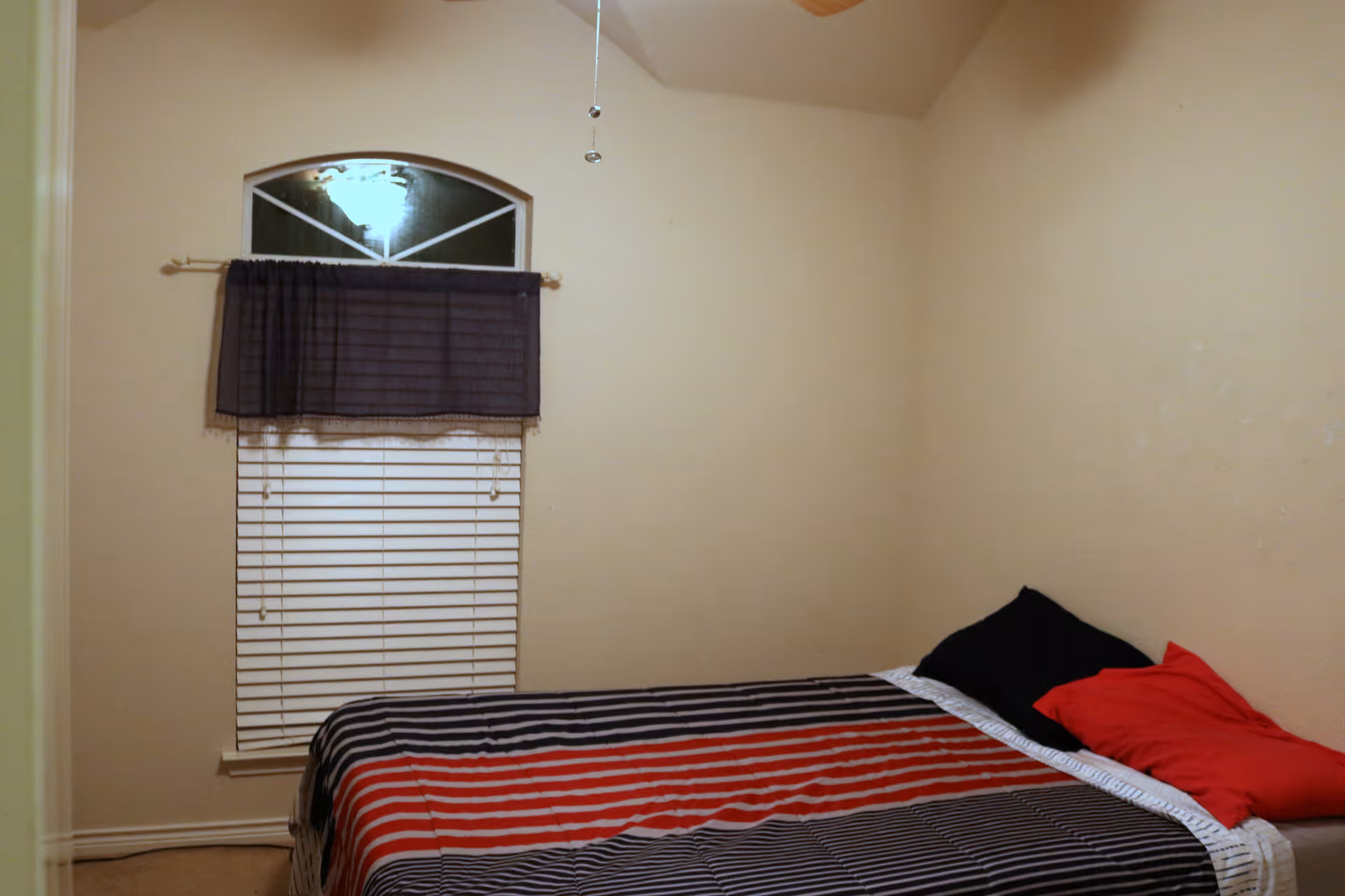 A small bedroom with beige walls, a bed with a striped black, red, and white bedspread, and two pillows, one black and one red. There is a window with white blinds and a dark curtain valance above it. A ceiling fan pull chain is visible hanging from the ceiling.