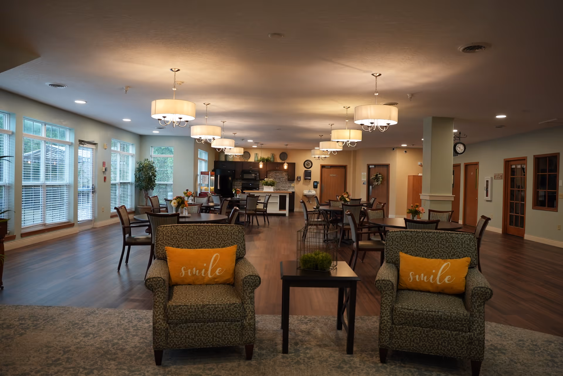 Spacious assisted-living common area with armchairs, dining tables, pendant lights and a kitchen area in the background.