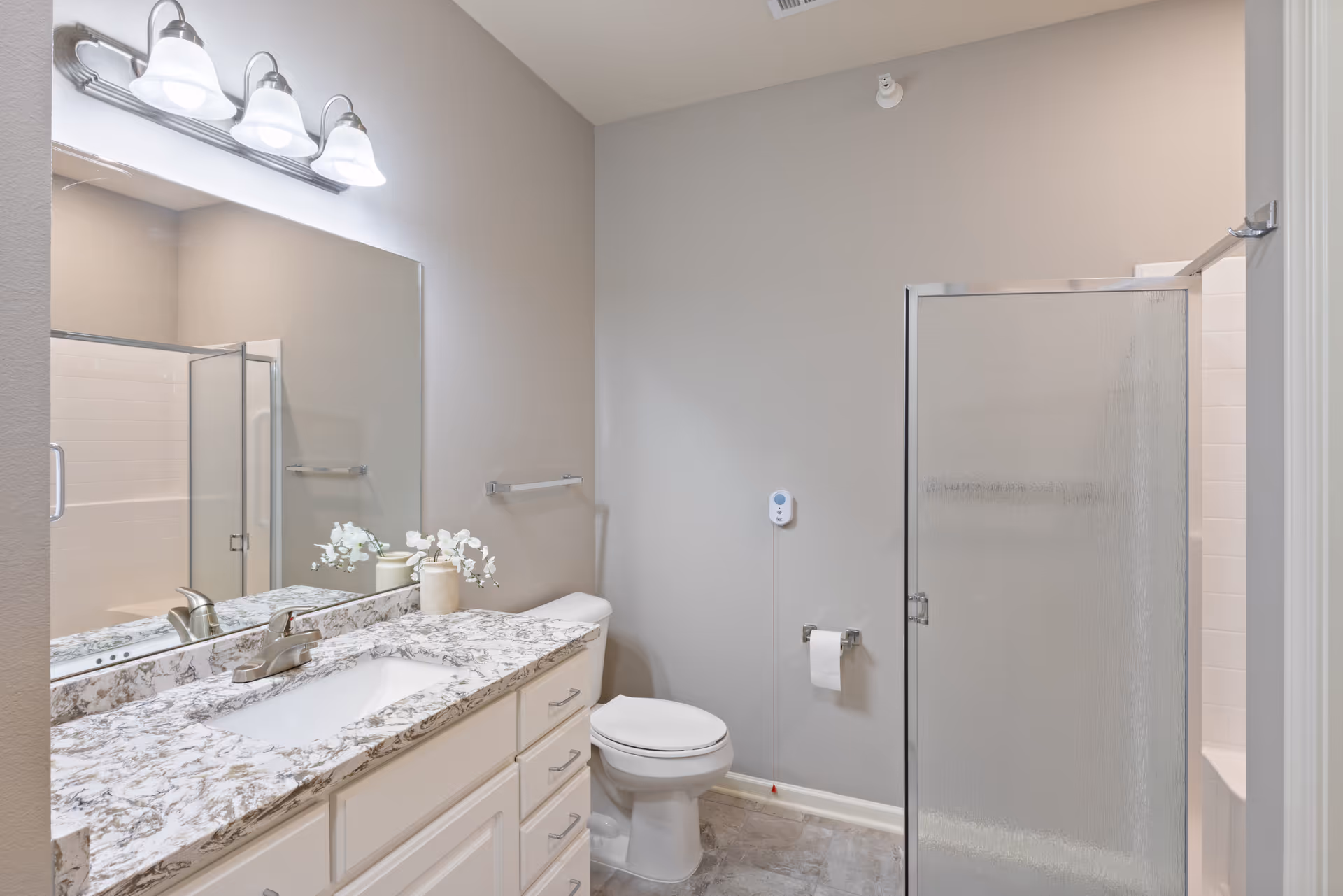 A clean and modern bathroom featuring a white vanity with a marble countertop, a large mirror above the sink, a toilet, and a glass-enclosed shower with textured glass. The walls are painted light gray and the floor has gray tiles. There is a small decorative plant on the countertop and a wall-mounted light fixture with three lights above the mirror.