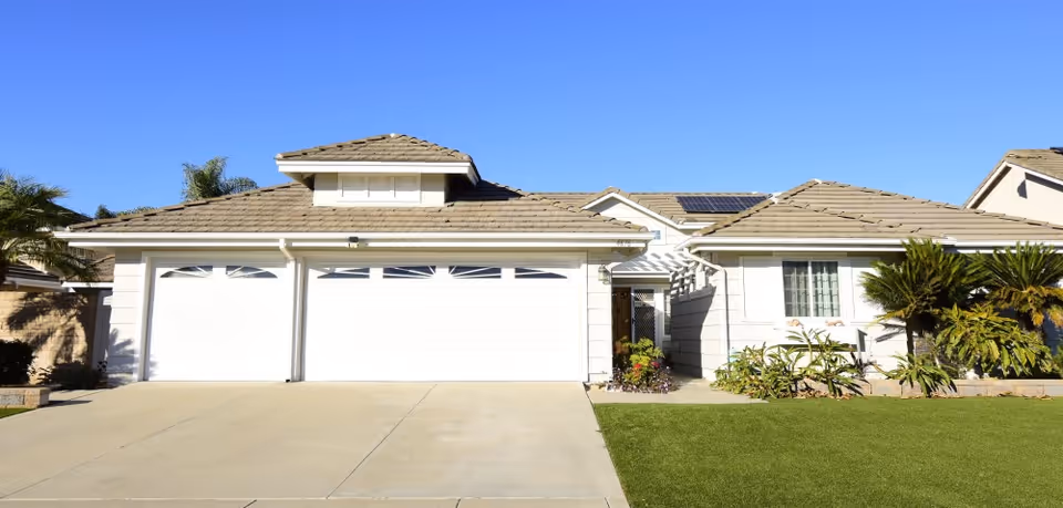 Front exterior view of a single-story house with a three-car garage, beige roof tiles, white garage doors, and a well-maintained lawn with some plants and shrubs under a clear blue sky.