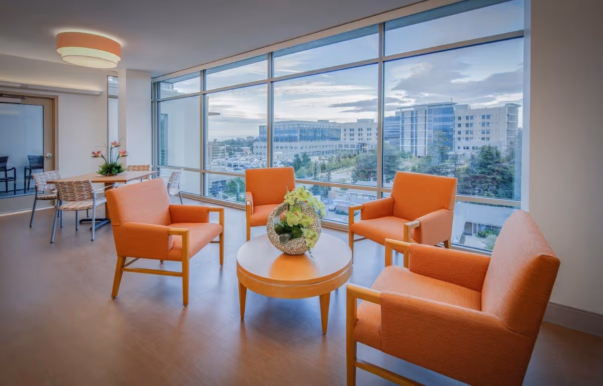 A bright seating area in a senior living facility with four orange armchairs arranged around a round wooden table with a decorative plant centerpiece. Large floor-to-ceiling windows provide a view of nearby buildings and trees outside. In the background, there is a small dining table with chairs and a modern light fixture on the ceiling.