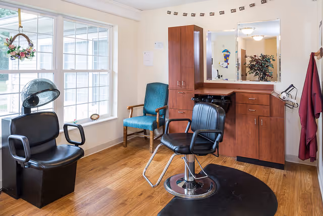 Interior view of a salon area in a senior living facility with a black salon chair on a black mat, a hair dryer chair near a large window, a teal upholstered chair, wooden cabinets with a mirror above, and a burgundy cape hanging on the wall.