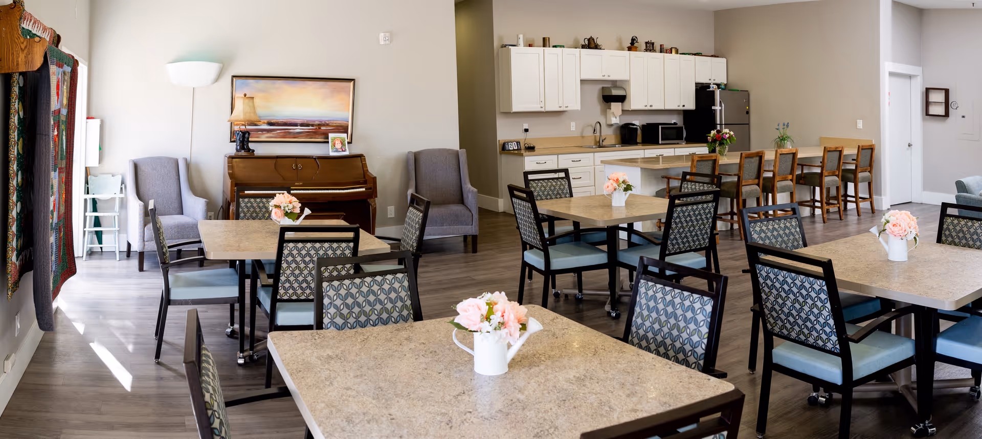 A bright and spacious common area in a senior living facility featuring multiple square tables with patterned chairs, each table decorated with a small vase of pink flowers. In the background, there is a kitchen area with white cabinets, a refrigerator, microwave, and a counter with several wooden bar stools. To the left, there is a piano against the wall flanked by two gray armchairs, a floor lamp, and a colorful quilt hanging on the wall. The room has light-colored walls and wood flooring.