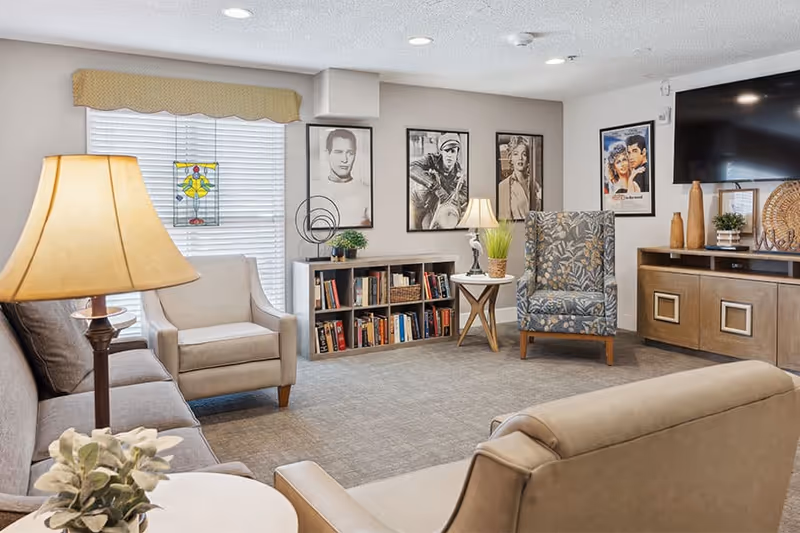 A cozy living room area in a senior living facility with beige and patterned armchairs, a sofa, a floor lamp, a small bookshelf filled with books, framed black and white portraits on the wall, a flat-screen TV mounted on the wall, and decorative plants and vases on a wooden cabinet.