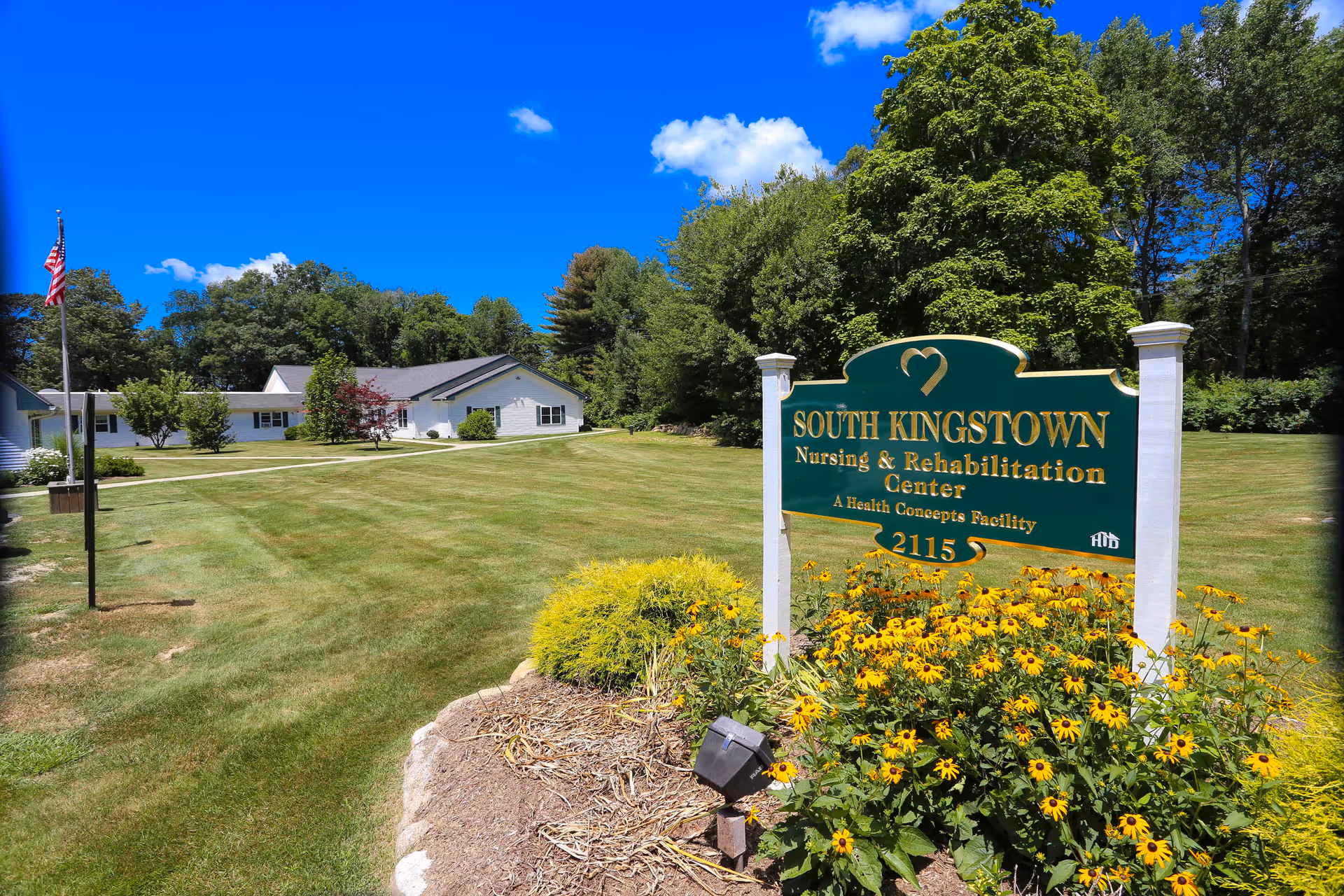 Entrance sign for South Kingstown Nursing & Rehabilitation Center on a manicured lawn with flowers, nearby buildings, an American flag, and trees under a blue sky.