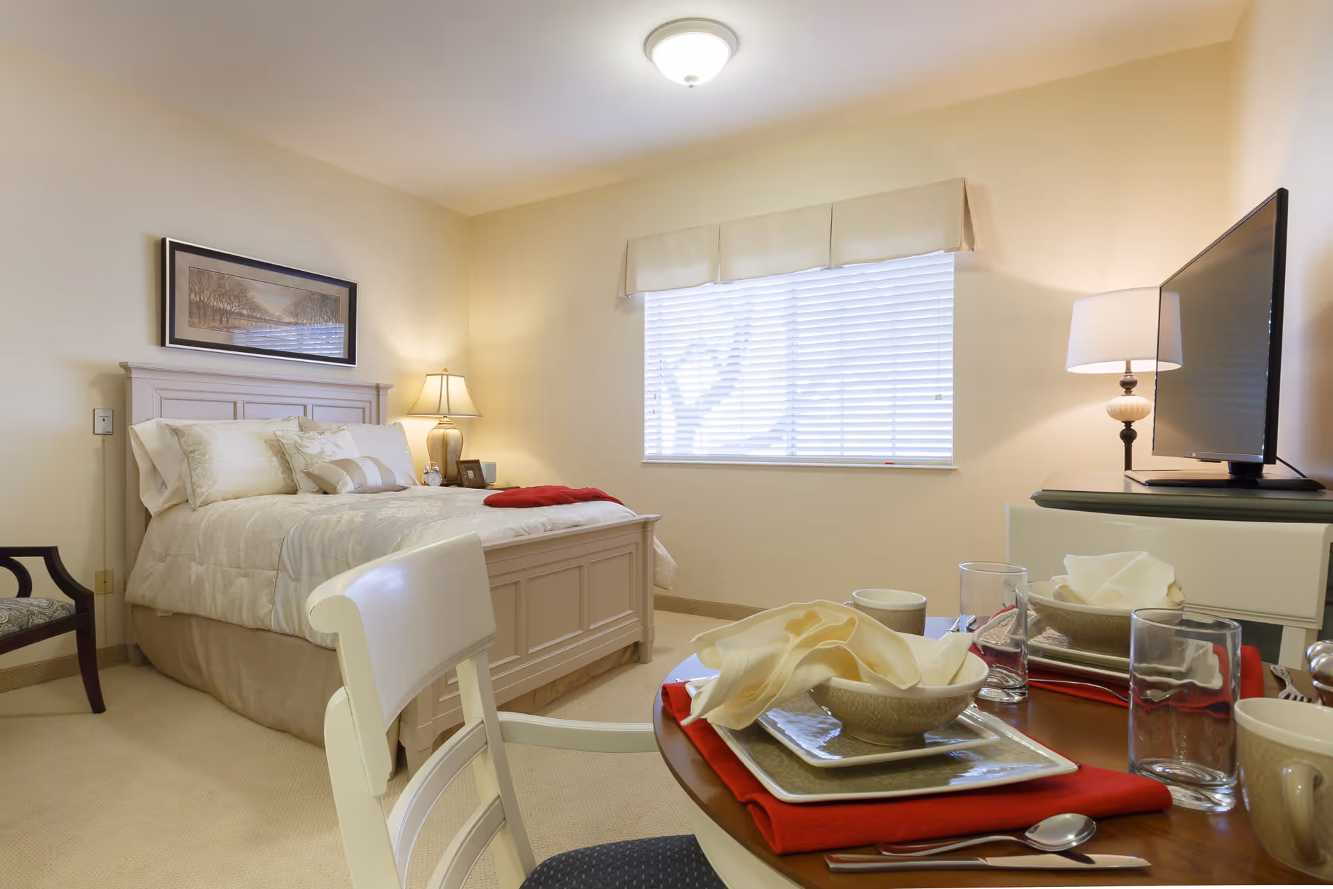 A cozy bedroom with a neatly made bed featuring cream-colored bedding and pillows. A bedside table with a lamp and picture frame is next to the bed. A window with blinds and a valance lets in natural light. In the foreground, there is a small dining table set with plates, bowls, glasses, and napkins on red placemats. A television and another lamp are on a dresser against the wall.
