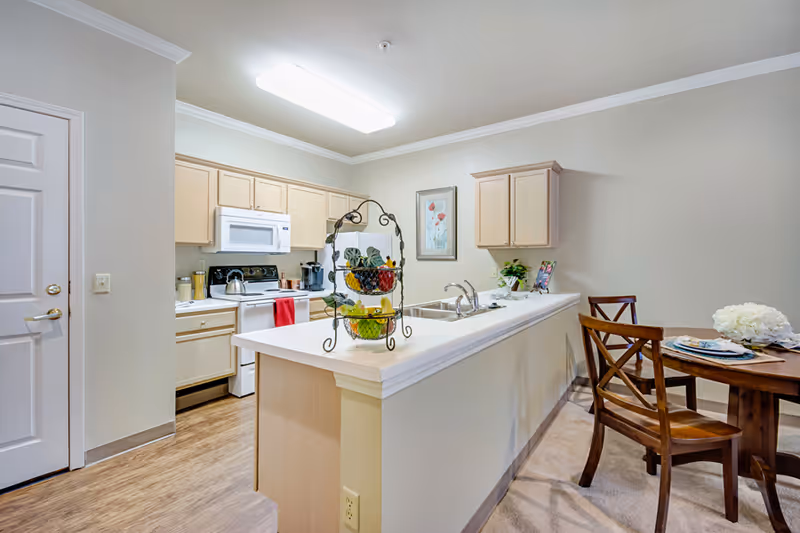 A bright kitchen area with light wood cabinets, a white microwave, stove, and dishwasher. A white countertop island with a double sink and a tiered fruit basket sits in the center. To the right, there is a small wooden dining table with two chairs, set with plates and a flower centerpiece. The floor is a combination of wood and carpet, and the walls are painted a light beige color.
