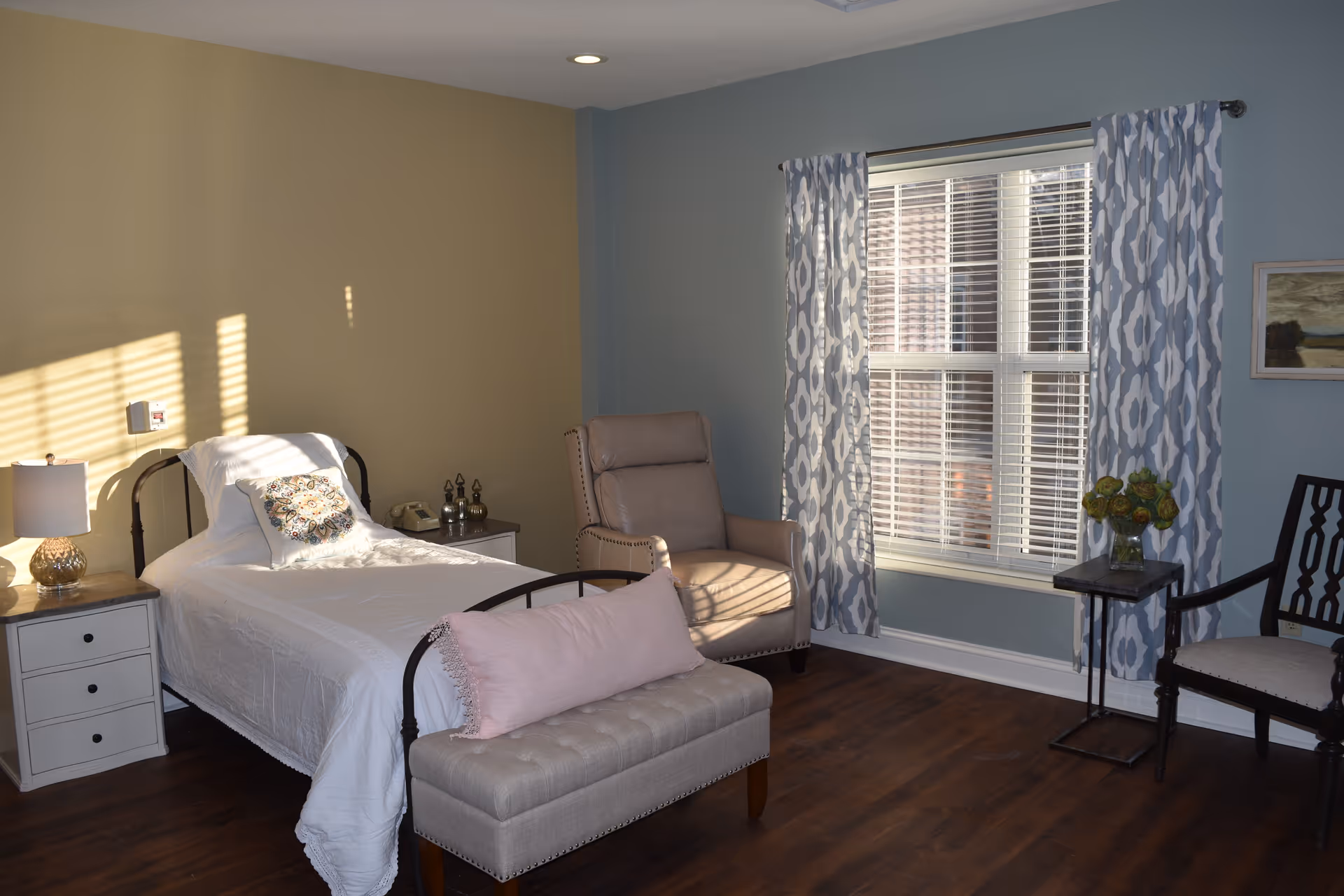 Sunlit bedroom with a single metal-framed bed, nightstands, an armchair and a window with patterned curtains.