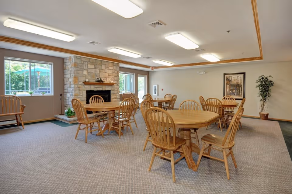Bright community dining room with round wooden tables and chairs, a stone fireplace, windows, and a potted plant.