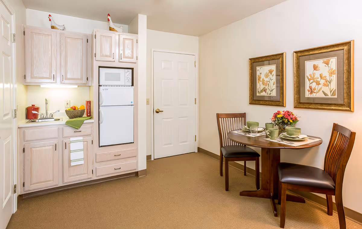 A small kitchen and dining area in a senior living facility. The kitchen features light wood cabinets, a sink, a microwave, and a refrigerator. On the right side, there is a round wooden dining table set for two with green dishes and a floral centerpiece. Two framed floral prints hang on the wall above the table.